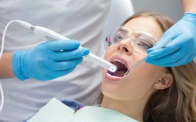 Lovely girl with opened mouth in patient bib and protective eyewear. Next to her there is a dentist in a white uniform with blue latex gloves. He diagnoses her teeth with a LED intraoral camera and a stem mouth mirror. Horizontal.